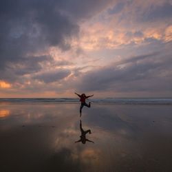 Woman jumping at beach against cloudy sky during sunset