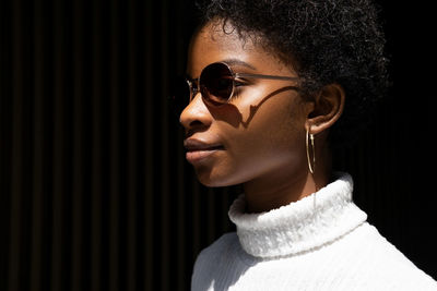 Side view of young african american woman in stylish sweater and sunglasses looking away while standing in bright sunlight against black background