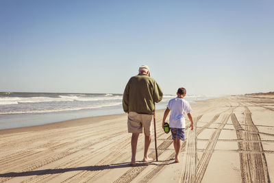 Rear view of grandfather and grandson walking on sand against clear sky