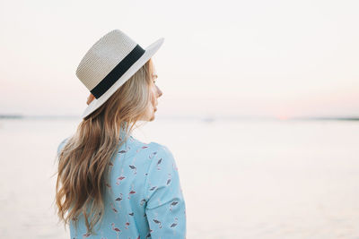 Beautiful blonde young woman in blue dress and straw hat on pier on sunset