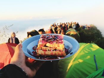 Cropped hand of person holding food in plate against people and sky