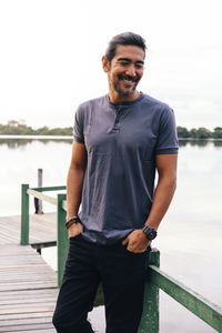 Smiling mature man with hands in pockets standing on pier against lake