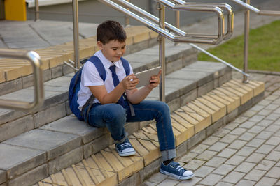 Schoolboy in a white shirt with a blue jacket sits on the stairs and plays with a gray tablet
