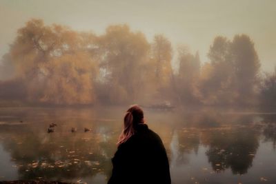 Woman standing by lake against trees during foggy weather
