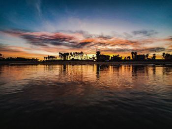 Silhouette buildings by lake against sky during sunset