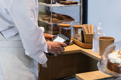 Side view of crop anonymous seller using pos terminal for payment while standing at counter in bakehouse with vegan desserts