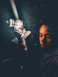 Close-up of woman looking away sitting by illuminated light with butterflies at night