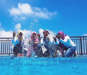 Low angle view of happy friends splashing water at poolside against blue sky during sunny day