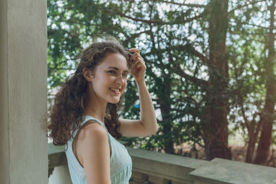 Beautiful curly hair young woman in light blue simple dress on balcony, park trees background