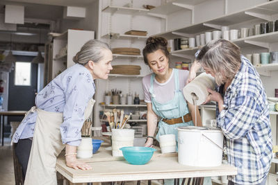 Senior potter and young employee looking at mature colleague pouring clay from vase in strainer on bucket at workshop