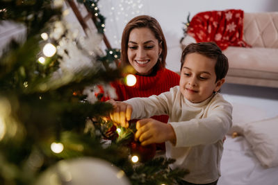 Cheerful festive family of woman with small child son decorating christmas tree with lights and toys