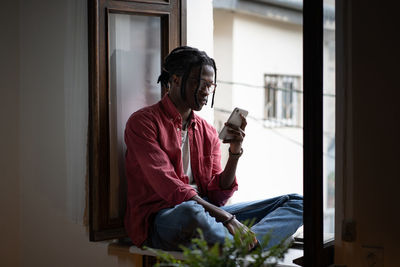 Young african guy sitting on windowsill freelancing with smartphone from home