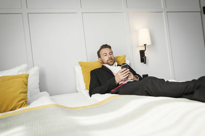 Businessman lying on bed while listening to music from mobile phone in hotel room