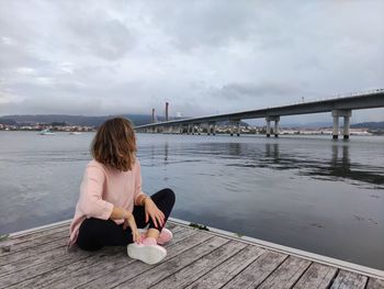 Rear view of woman sitting on pier over bridge against sky