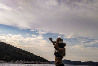 Side view of woman wearing hat and bikini standing at beach against cloudy sky