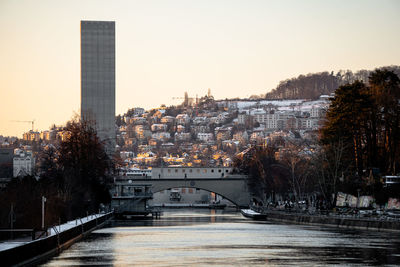 Bridge over river by buildings in city against sky at sunset