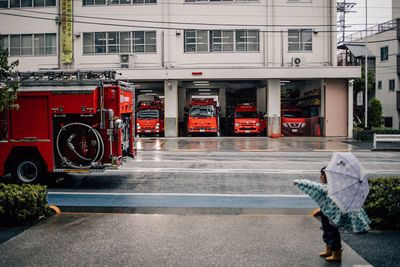 A three year old child pointing at a running  firetruck in front of a fire station.