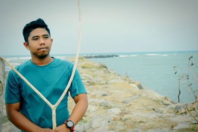 Thoughtful young man looking away with stick at beach against sky