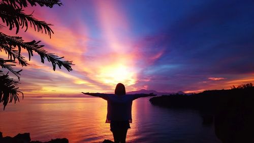 Silhouette woman with arms outstretched standing on shore against orange sky