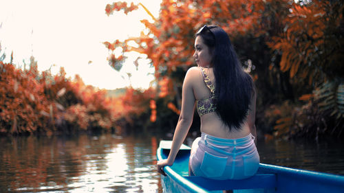 Young woman looking away while sitting by lake against trees