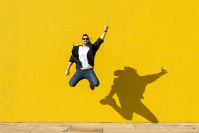 Young man with sunglasses jumping in front of a yellow wall.