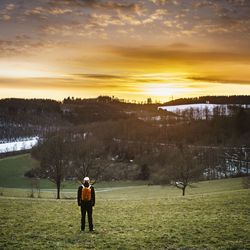 Rear view of young man standing on grassy field against orange sky during winter