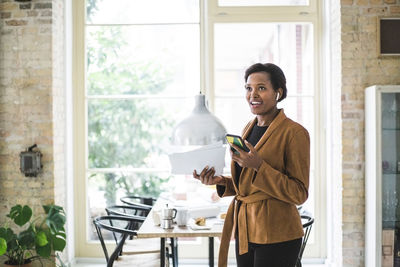 Smiling female entrepreneur using phone while standing by table at home
