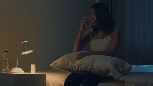 Young woman drinking from glass while sitting on bed