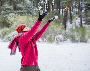 Cheerful young woman throwing snow mid air while standing on snow covered land