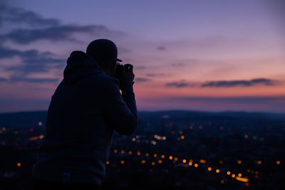 Man photographing illuminated cityscape against sky during sunset