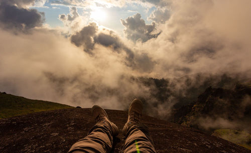 Low section of person sitting on mountain against cloudy sky during sunset