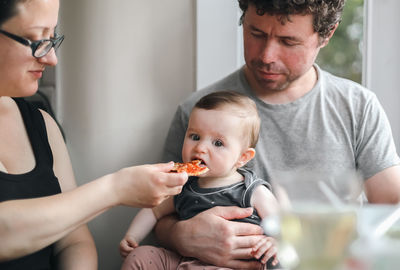 Portrait of a caucasian girl sitting in her father's arms, and her mother feeds her a piece of pizza