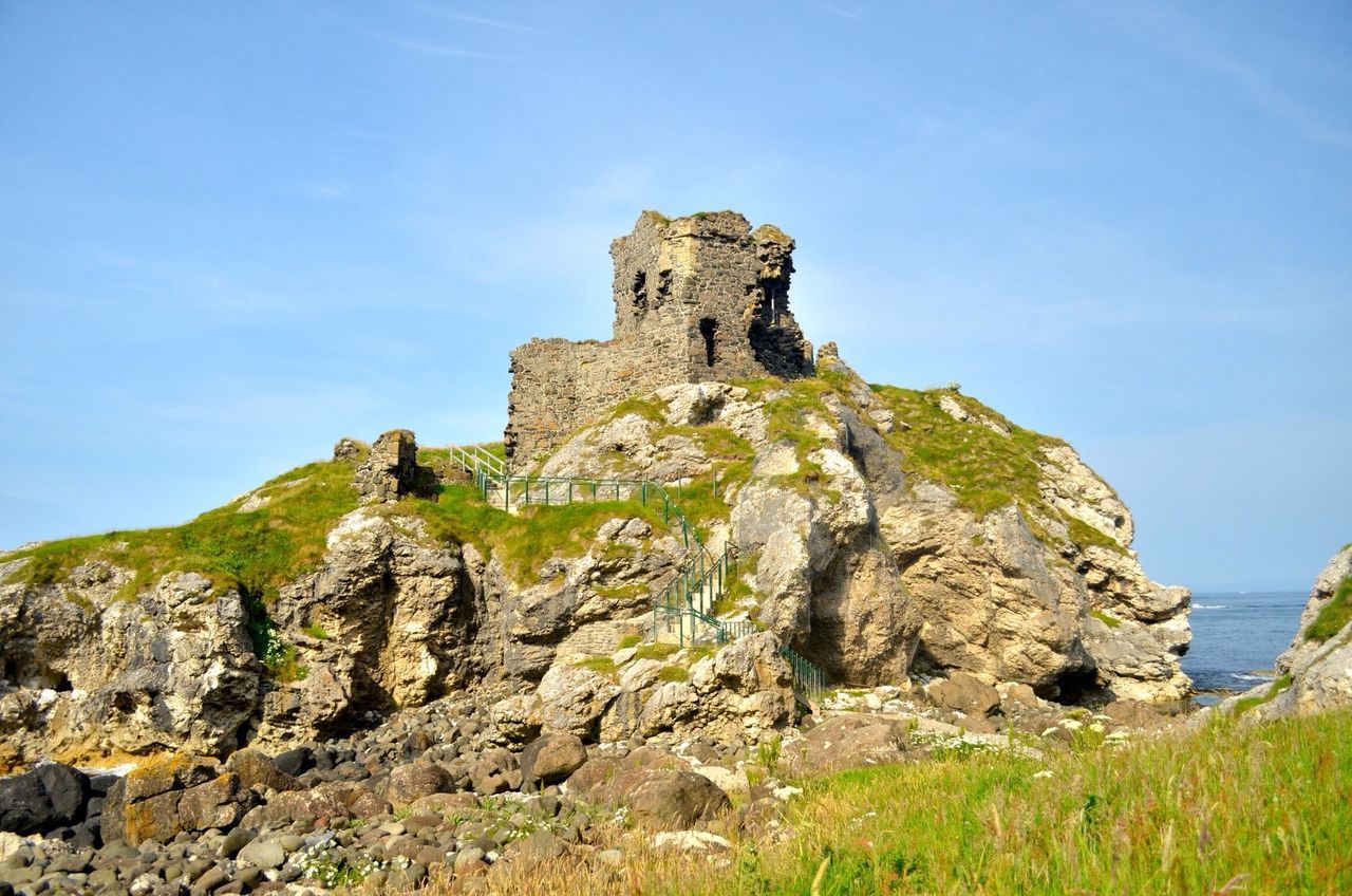 sky, rock formation, built structure, history, architecture, grass, old ruin, tranquility, rock - object, blue, cliff, landscape, low angle view, ancient, tranquil scene, nature, travel destinations, the past, building exterior, famous place