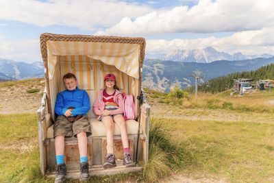 Full length of happy siblings sitting in hooded beach chair on grassy field against sky