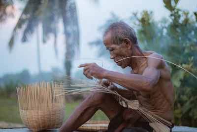 Shirtless senior man making wicker basket while sitting at farm