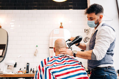 Anonymous male stylist in mask using hairdryer while touching bald head of man in striped cloth in beauty salon