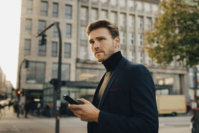 Contemplating businessman with smart phone looking away while standing against building in city