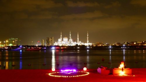 Illuminated building by river against sky at night