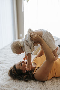 Cheerful mother holding toddler son and playing while lying down at home