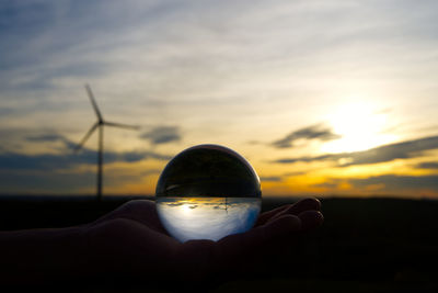 Hand holding crystal ball against sky during sunset