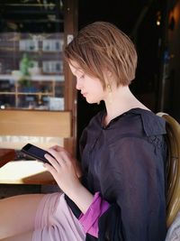 Side view of beautiful young woman using mobile phone while sitting on chair at home