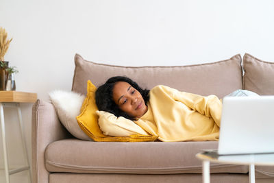 Young african-american female watching movies on laptop while lying on sofa in living room