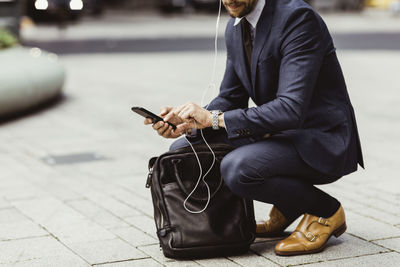 Low section of male entrepreneur using smart phone through in-ear headphones while crouching on street