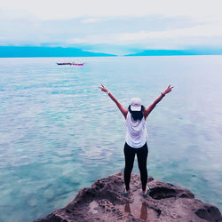 Rear view of young woman with arms raised standing on rock by sea against cloudy sky