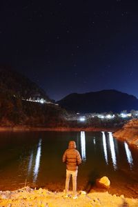 Rear view of man standing by lake against sky at night