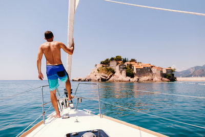 Full length rear view of shirtless man standing on yacht sailing on sea