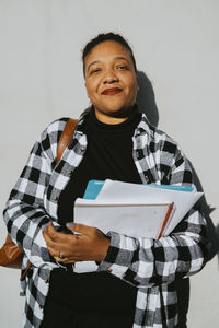 Portrait of smiling mature woman holding book standing against wall on sunny day