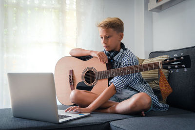 Full length of boy using laptop while learning guitar on sofa at home