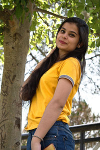 Low angle portrait of young woman standing against tree trunk