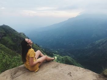 High angle view of woman laughing while sitting on rock against mountains and sky